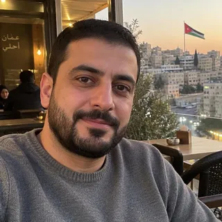 صورة شخصية لمازن الحوراني، رجل في منتصف الثلاثينيات بشعر أسود قصير ولحية مشذبة يرتدي كنزة رمادية في مقهى بعمّان